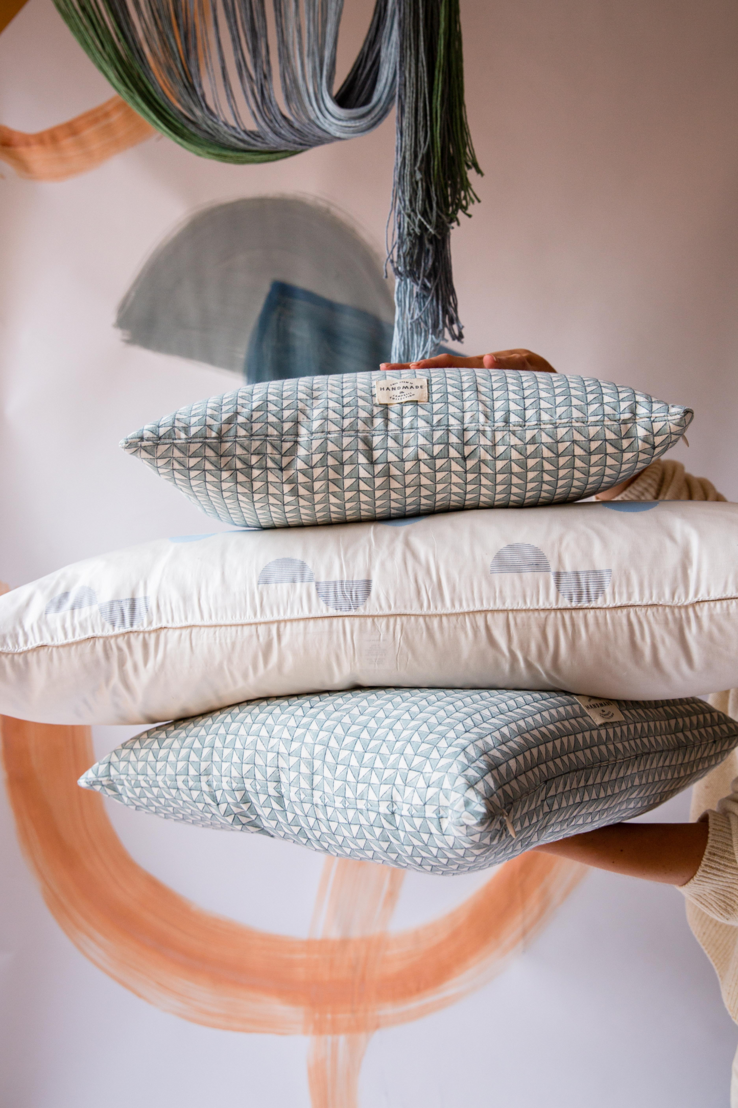 Someone holding a pile of three cushions. The top and bottom cushion are blue with a triangle pattern on and the middle cushion is cream with blue spots on