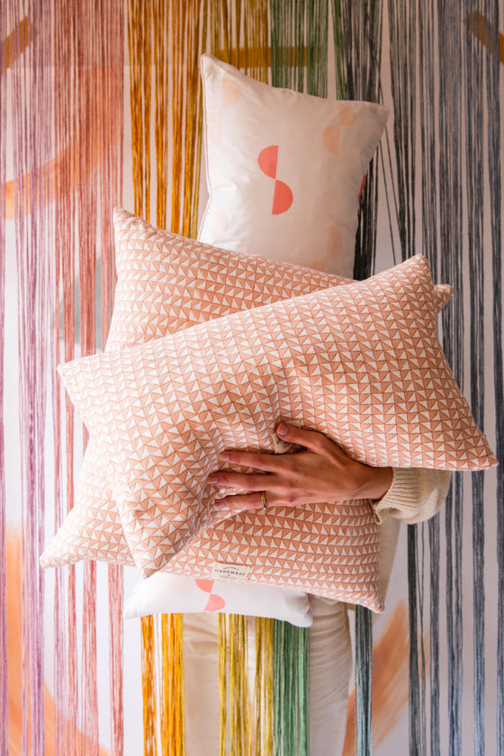 Someone holding a pile of three cushions. The front two cushion are pink with a triangle pattern on and the back cushion is cream with pink spots on
